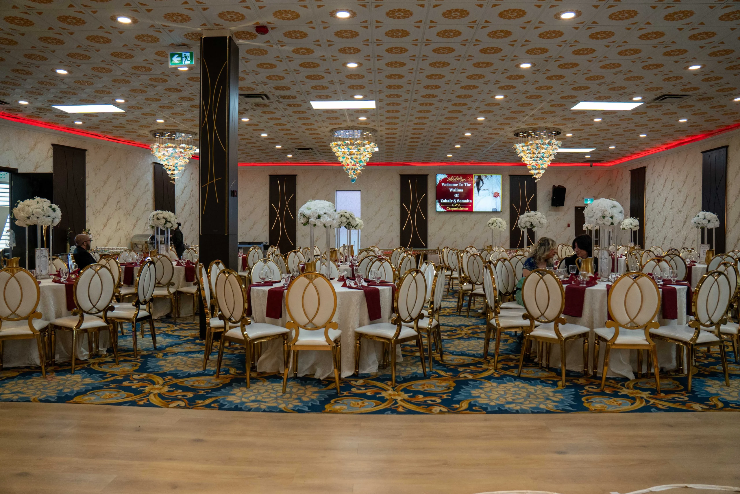 Elegant small wedding reception with floral centrepieces, decorated dining tables and gold chairs in a banquet hall.